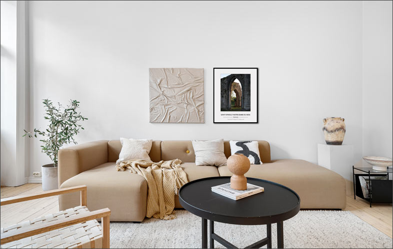 L’image met en scène un intérieur mêlant authenticité et modernité, où les matériaux bruts — notamment la pierre — occupent une place centrale. Cet équilibre entre rusticité et lignes contemporaines est caractéristique des styles actuels qui cherchent à conserver le charme ancien tout en apportant confort et sobriété.
L’espace dégage une atmosphère chaleureuse et élégante, avec une décoration volontairement épurée qui laisse s’exprimer les textures naturelles et la lumière. Le mobilier moderne contraste subtilement avec le caractère plus traditionnel du lieu, créant un ensemble harmonieux et intemporel.
Au mur, une photographie encadrée attire le regard : elle représente les ruines de l’abbaye de Saint-Évroult-Notre-Dame-du-Bois au crépuscule. Cette image patrimoniale, aux tonalités douces et légèrement dramatiques, renforce le lien entre passé et présent, tout en apportant une profondeur visuelle et une dimension culturelle à la pièce.