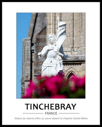 vue de la statue enti�rement blanche de Jeanne d'Arc dont le bas est cach� par des fleurs fuschia d'aspect flou en avant-plan. A l'arri�re, on voit une partie gauche de la chapelle et le ciel bleu