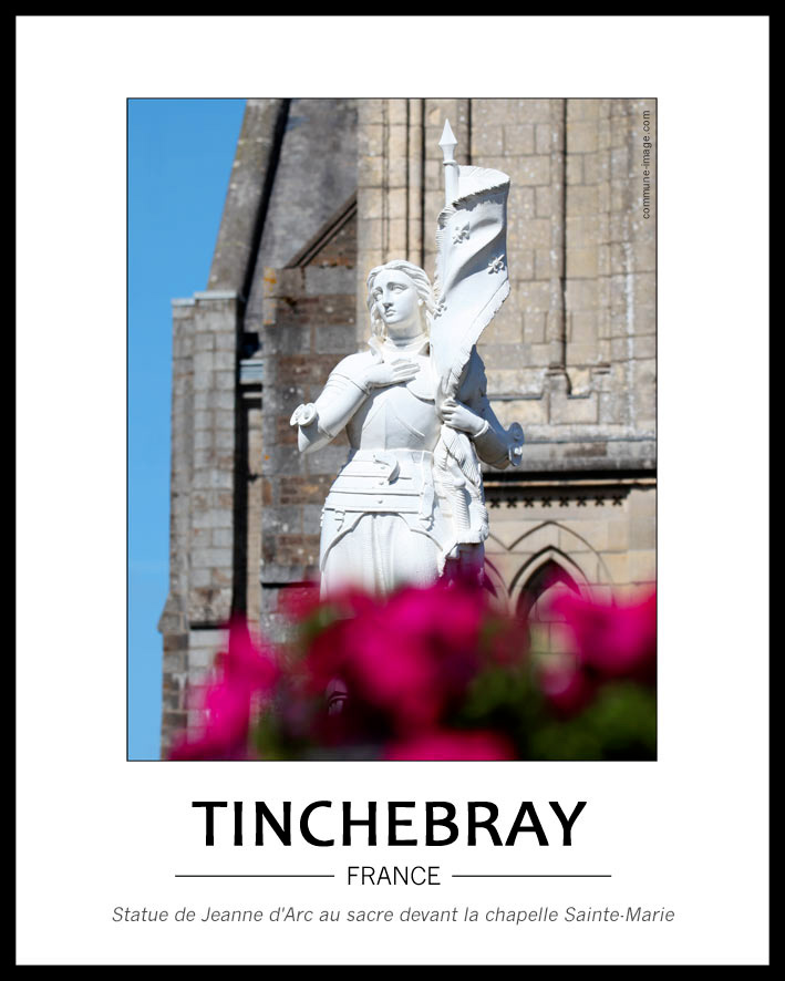 Cliquez pour agrandir vue de la statue entièrement blanche de Jeanne d'Arc dont le bas est caché par des fleurs fuschia d'aspect flou en avant-plan. A l'arrière, on voit une partie gauche de la chapelle et le ciel bleu